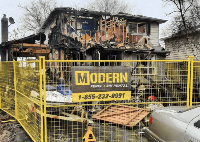 Yellow temporary fence rentals surrounding a burned down house. The fence has a Modern Fence Rental sign on it.